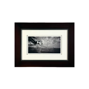 Framed black-and-white photograph of a seaside town along a rocky shore with a small sailboat on the water.