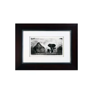 Black-and-white photo of elephants near thatched huts in a misty forest, framed by a dark wooden frame.