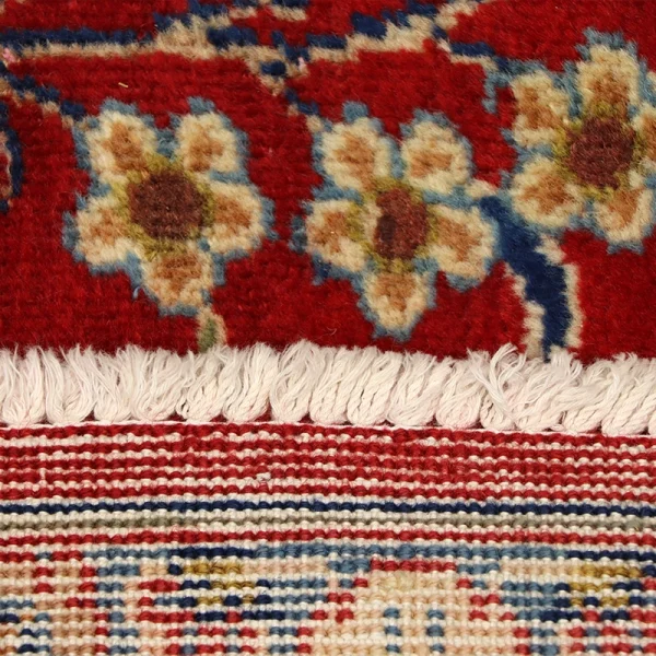 Close-up of a red rug edge with white fringe and floral cream, gold, and blue patterns.