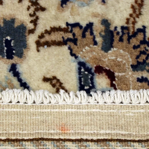 Close-up of a patterned rug showing beige, blue, and brown motifs with a white fringe along the edge.