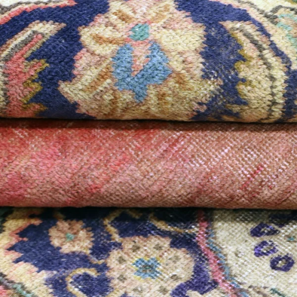 Rolled, patterned area rugs stacked on top of each other, showing ornate blue, beige, and pink designs.