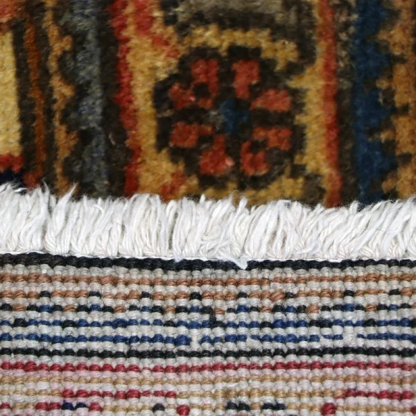 Close-up of two woven rugs: a fringed white edge sits atop a colorful patterned rug with a red floral motif in the center.