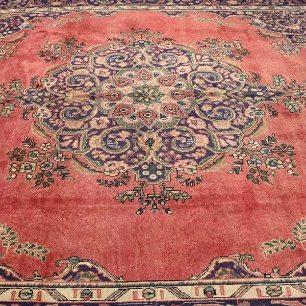 Persian-style area rug with a pinkish-red background and a detailed central medallion in purple, blue, and cream hues. Ornate oriental rug laid flat, featuring a floral border and a vibrant, multicolored central motif on a coral field.