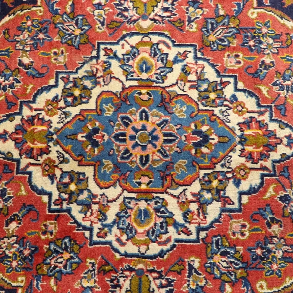 Intricate patterned rug with a central blue medallion and red surroundings, featuring floral motifs in white, yellow, and pink hues
